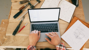 Builder using a laptop with construction plans and tools on a workbench, representing construction ERP software connecting field activity with financial data