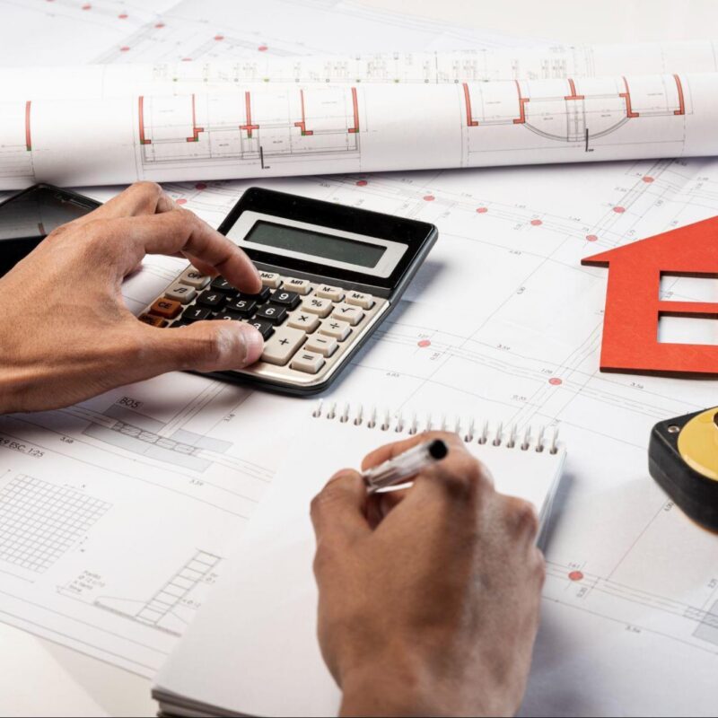 Person using a calculator and taking notes while reviewing architectural blueprints, with a smartphone, measuring tape, and house-shaped model placed on a desk.