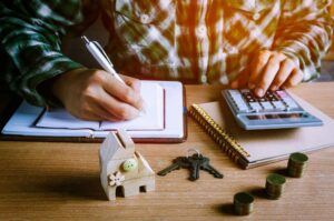 Person writing in a notebook while using a calculator at a desk, with a small wooden house model, keys, coins, and office supplies nearby.